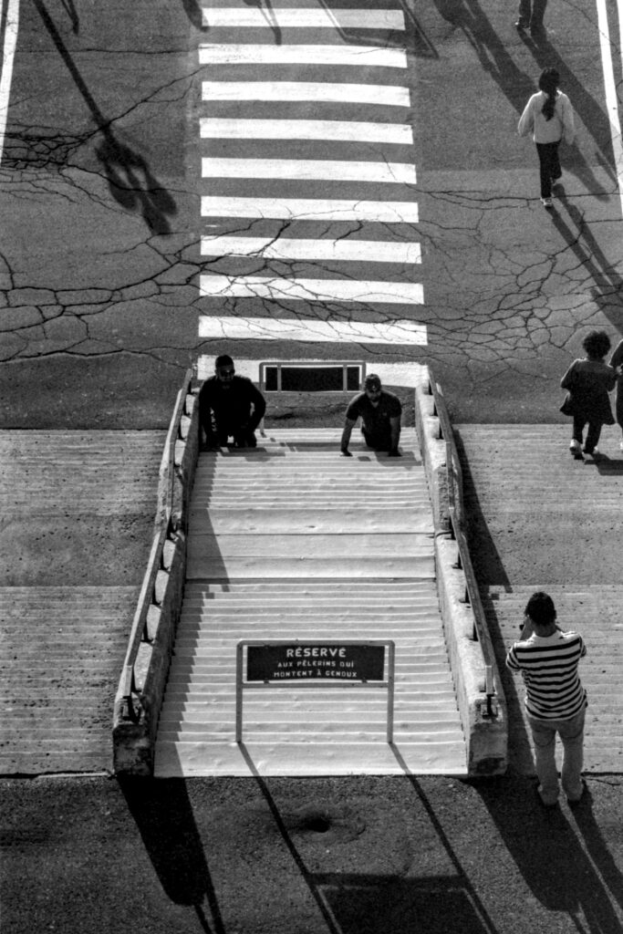 A view fron the top. At the bottom some stairs, with signs saying they are reserved for pilgrims climbing on the knees. Two persons are doing so. Further up a crosswalk. The asphalt shows visible cracks. People a walking down the stairs to the side. The sun cast shade towards the bottom right of the frame.

The picture is in black and white.